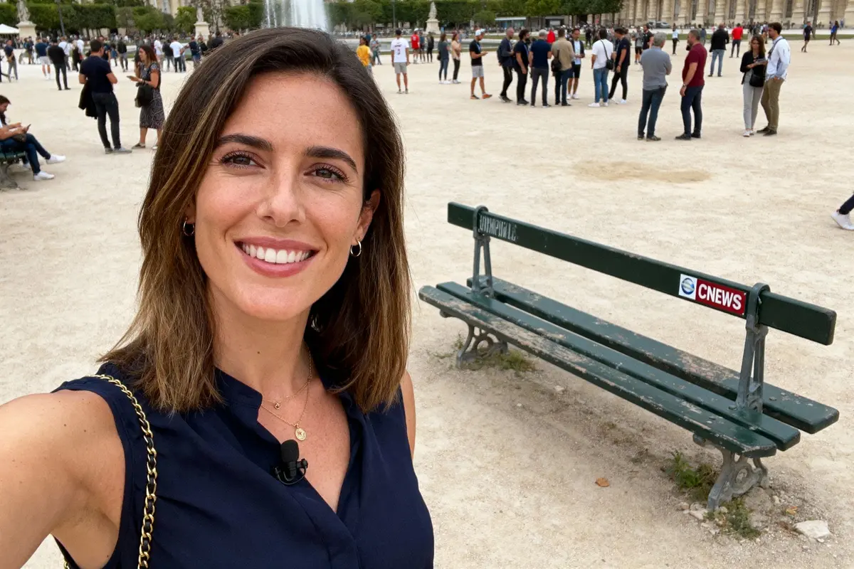 Femme souriante prenant un selfie devant un banc, avec des gens en arrière-plan dans un parc.