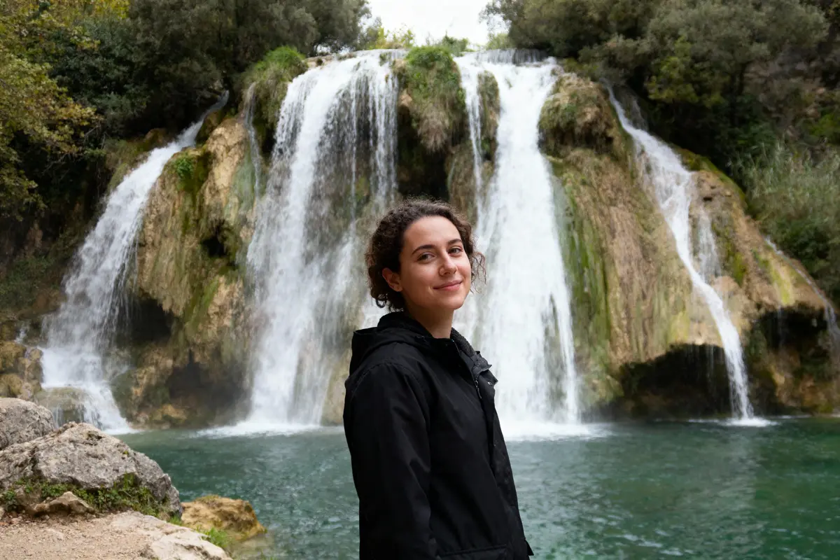 Une personne souriante se tient devant une grande cascade entourée de verdure et d'eau claire.