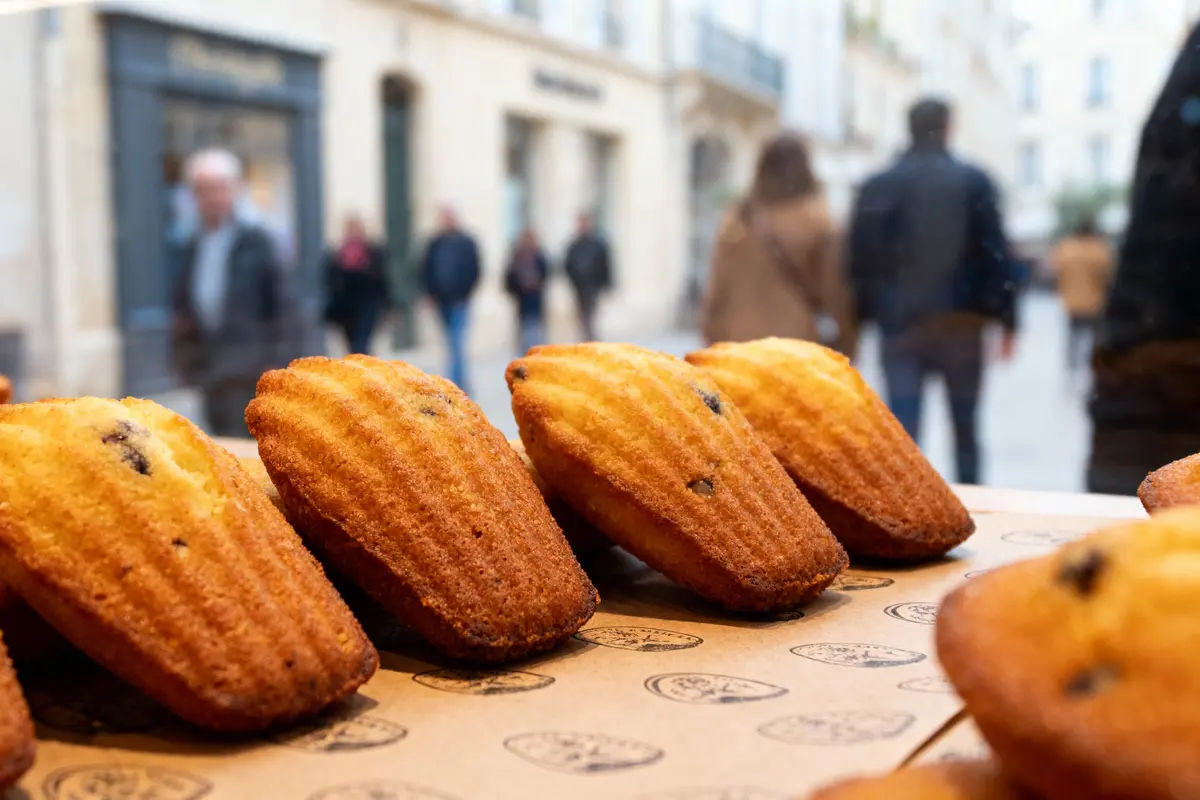 Madeleines dorées en vitrine d'une boulangerie avec des passants flous en arrière-plan.