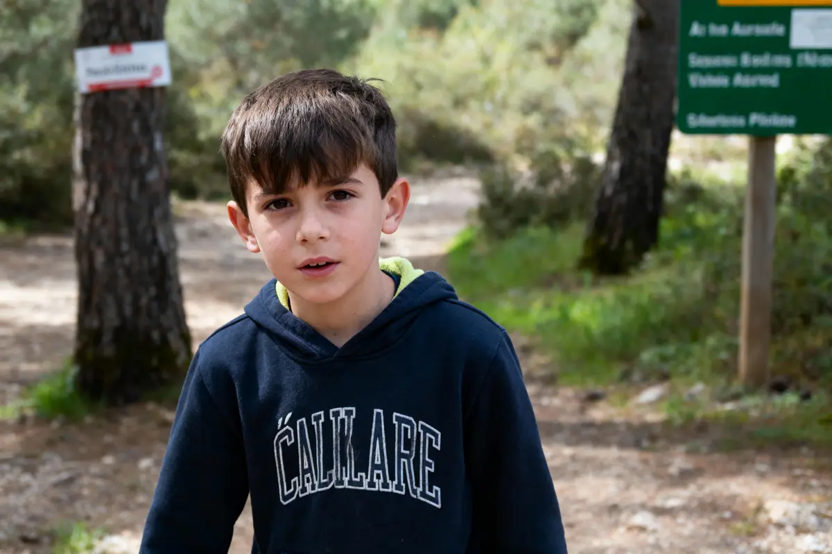 Jeune garçon en sweat à capuche bleu, debout sur un chemin forestier avec panneaux en arrière-plan.
