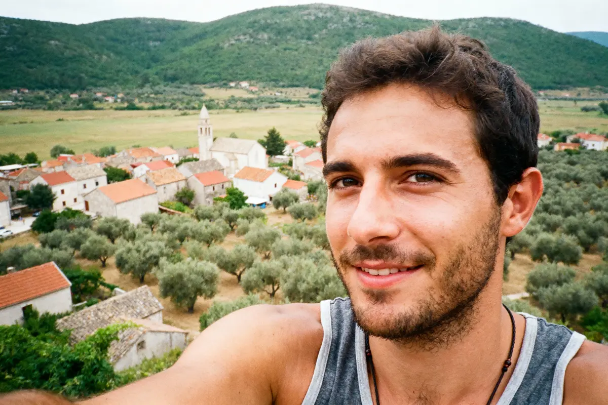 Homme souriant en selfie devant village pittoresque, oliviers et collines verdoyantes en arrière-plan.