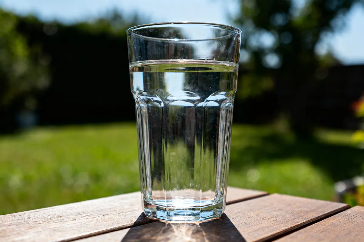 Verre d'eau sur une table en bois, jardin flou en arrière-plan, journée ensoleillée.