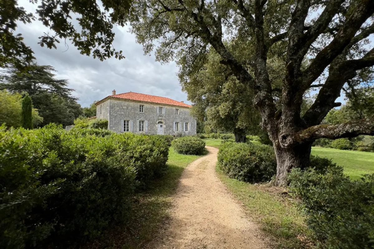Maison en pierre, toit en tuiles, chemin sinueux, arbres et buissons autour, ciel nuageux, ambiance campagne.