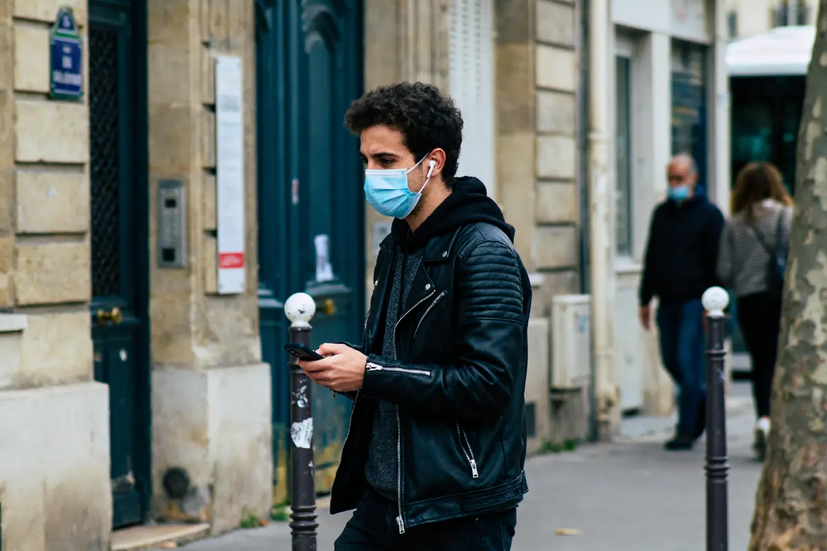 Homme portant un masque et des écouteurs, marchant dans une rue urbaine, utilisant un smartphone.