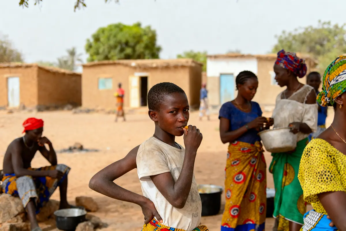 Un garçon mange, entouré de femmes occupant différentes tâches, dans un village africain à l'ambiance animée.