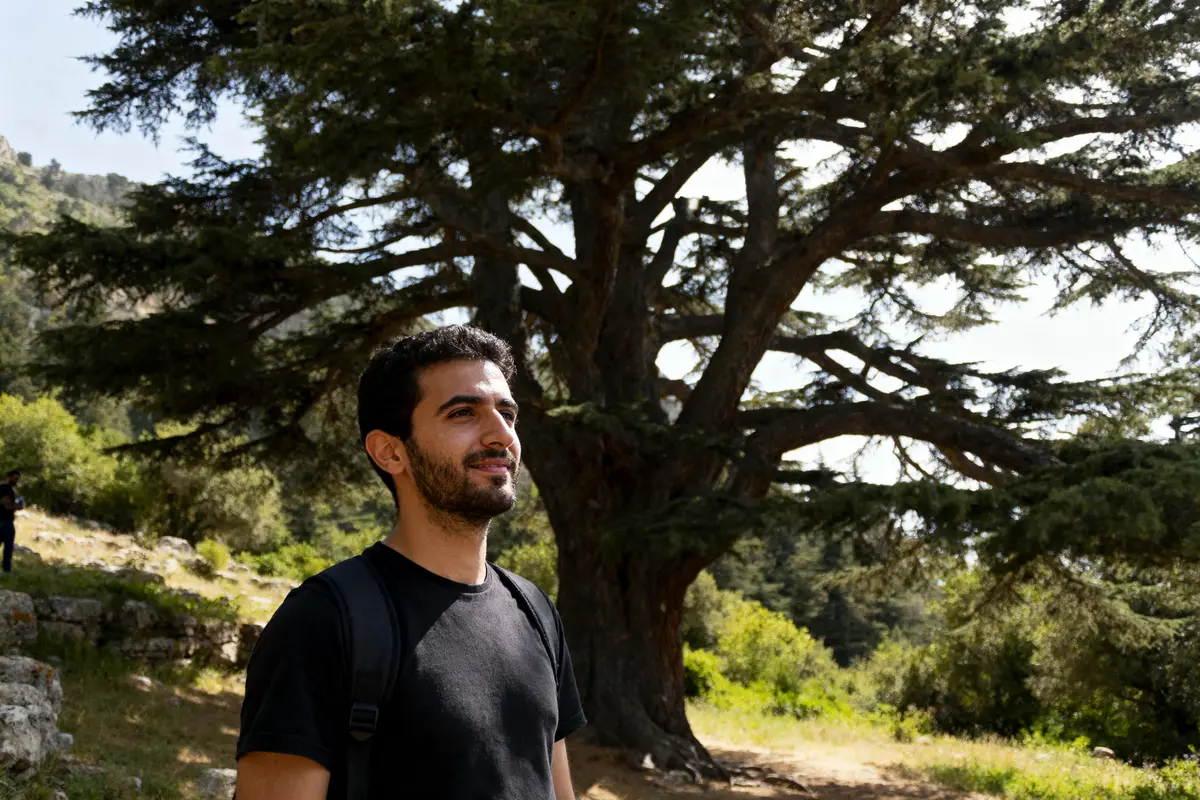 Un homme souriant, en plein air, debout devant un grand cèdre, par une journée ensoleillée.