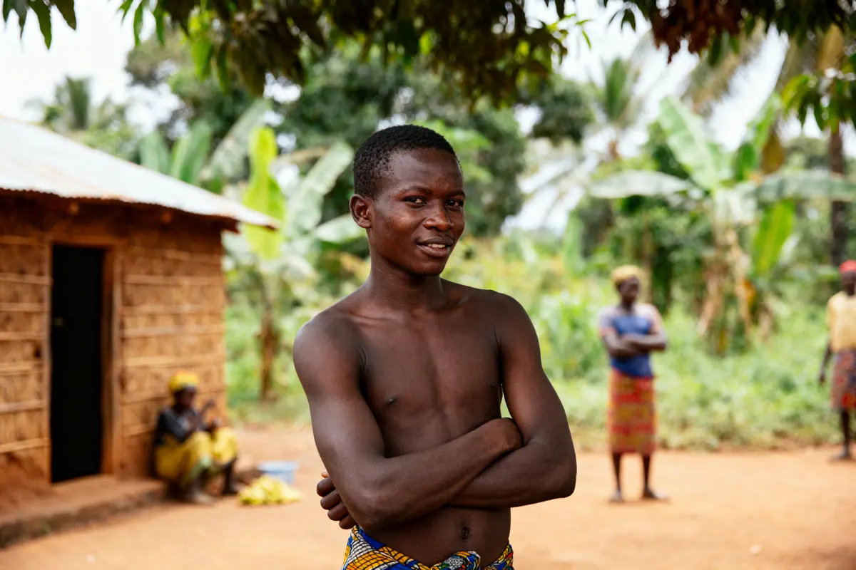 Un homme torse nu pose devant une maison en terre, avec végétation en arrière-plan.