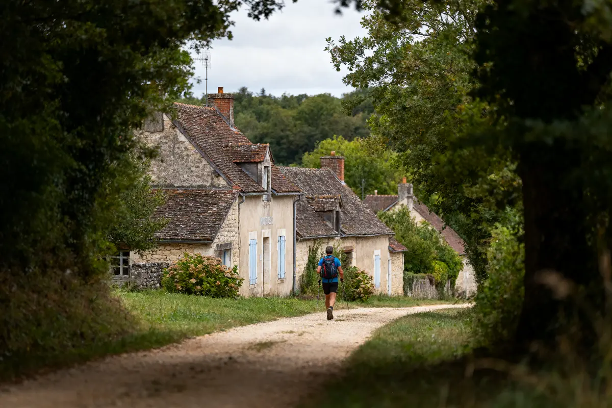 Personne marche dans un village rural, maisons en pierre, arbres entourant le chemin. Ambiance calme et naturelle.