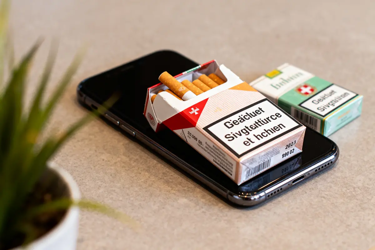 Paquet de cigarettes ouvert sur un smartphone, boîte fermée à côté, plante floue en arrière-plan.