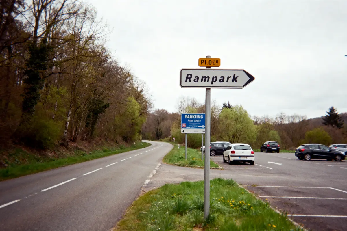 Route menant à un parking avec panneau "Rampark", entouré d'arbres et de voitures stationnées.
