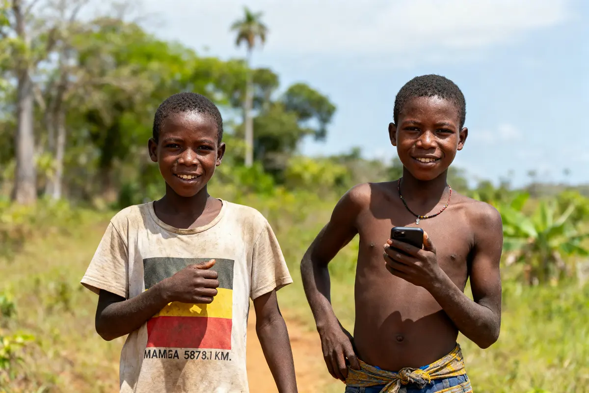 Deux garçons souriant, debout dehors, l'un tenant un téléphone portable, végétation verdoyante en arrière-plan.