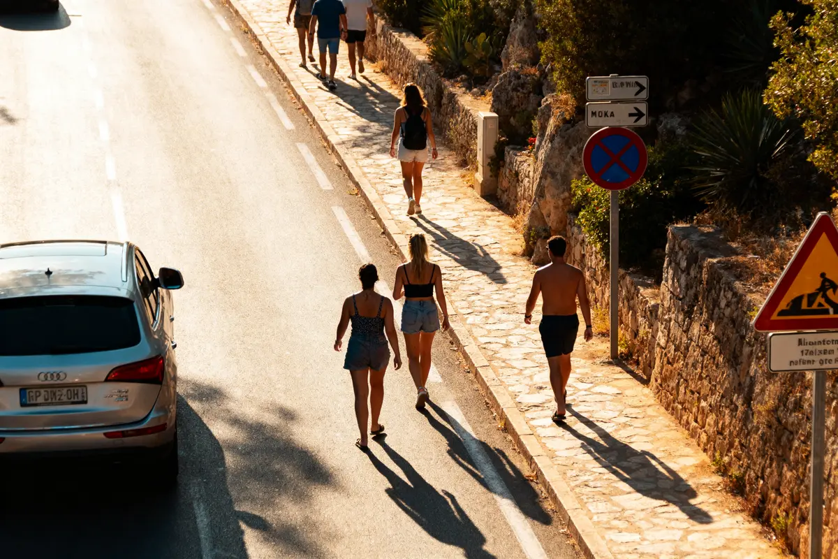 Personnes marchant sur un trottoir ensoleillé le long d'une route avec une voiture et des panneaux de signalisation.