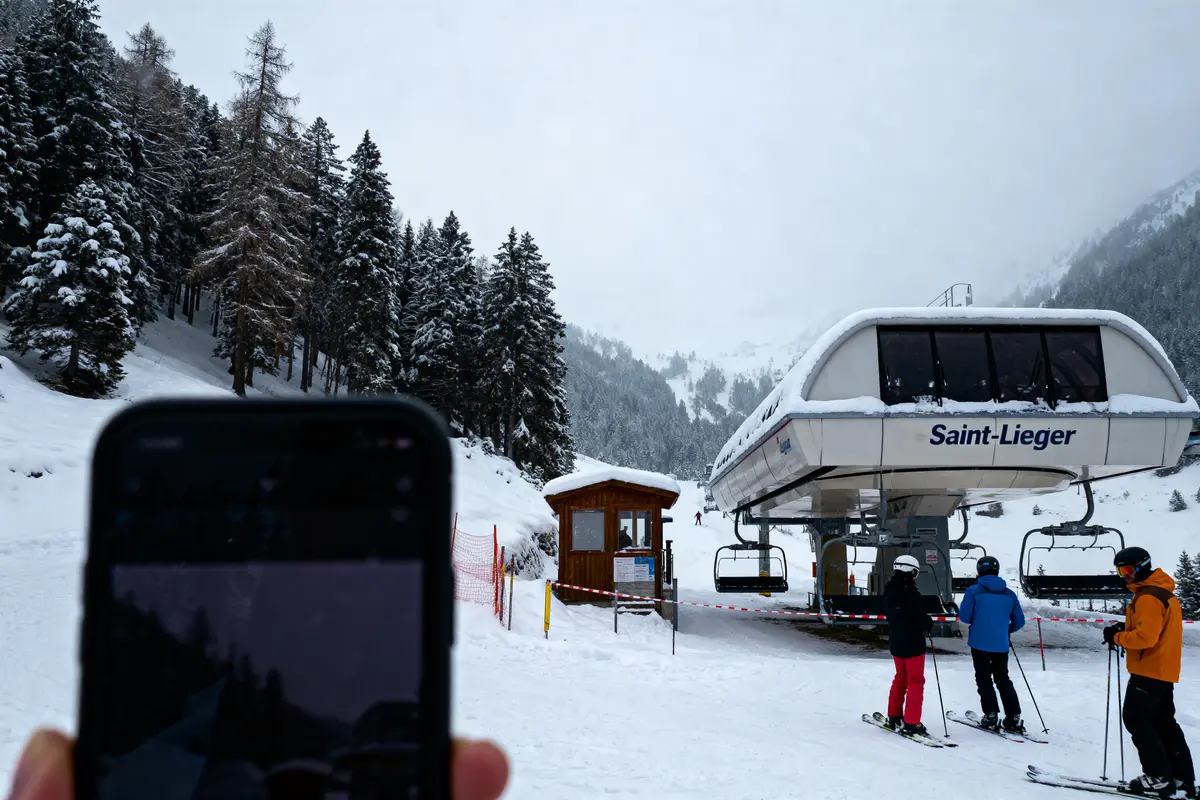Skieurs attendant un télésiège sous la neige, forêt enneigée à l'arrière-plan, téléphone en premier plan flou.